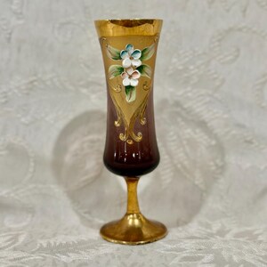 May include: A vintage cordial glass with a gold base and rim. The glass has a dark brown body with a gold floral design and hand-painted flowers in shades of white, blue, and pink. The stem and base are gold.