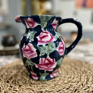 May include: A ceramic pitcher with a dark blue background and a floral pattern of pink roses and green leaves. The pitcher has a curved handle and a spout for pouring. It is sitting on a woven mat.