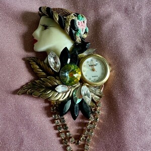 Puede incluir: Un broche dorado con un rostro de porcelana blanca con cabello negro y un adorno floral. El broche también incluye un reloj dorado con una esfera blanca y agujas negras, rodeado de diamantes de imitación y piedras verdes y negras.
