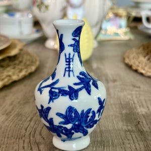 May include: A small, white porcelain vase with a blue and white floral design. The vase has a round body and a narrow neck. It is decorated with a floral pattern and Chinese characters.