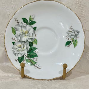 May include: White porcelain saucer with a gold rim and a floral design featuring white flowers and green leaves.