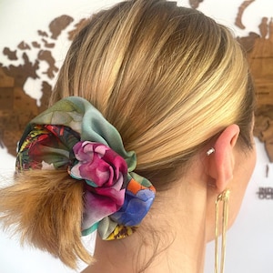 Puede incluir: Una goma para el pelo floral en tonos rosas, azules y verdes, atada alrededor de una coleta rubia. La goma para el pelo presenta un estampado botánico. Pendientes dorados cuelgan cerca de la oreja.