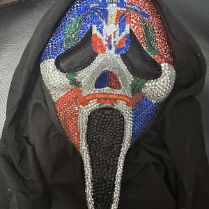 May include: A decorative mask with a Scream-style design, embellished with red, white, blue, and silver rhinestones. The mask features the Dominican Republic flag and the words "Dios, Patria, Libertad". The eye and mouth openings are black.