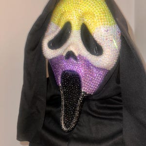 May include: A Ghostface mask covered in sparkling rhinestones featuring a yellow forehead, white eye areas, a purple nose and cheek section, and a black elongated mouth. The mask is attached to a black fabric hood that drapes around the sides.
