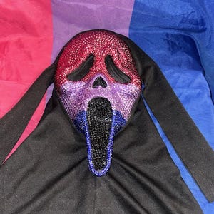May include: A bedazzled Scream mask in red, purple, and blue, with a black hood. The mask's design features a gradient of colors, with red at the top, transitioning to purple, and then blue at the bottom. The mask is set against a backdrop of pink, purple, and blue fabric.