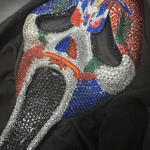 May include: A Scream mask covered in silver, red, blue, and green rhinestones. The mask has a black mesh eye area and a black chin. The top of the mask has text in white. The mask is resting on a black surface.
