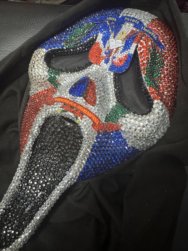May include: A Scream mask covered in silver, red, blue, and green rhinestones. The mask has a black mesh eye area and a black chin. The top of the mask has text in white. The mask is resting on a black surface.