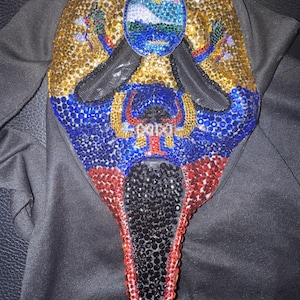 May include: A decorative mask covered in gold, blue, red, and black rhinestones. The mask features a central oval design with a landscape scene. The word "PAPA" is visible on the mask. The mask is on a black surface.