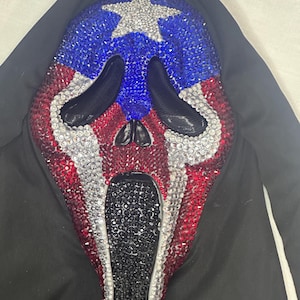 May include: A Scream mask with a patriotic design. The mask features a blue area with a silver star, red and white stripes, and is covered in sparkling embellishments. The mask is set against a black background.