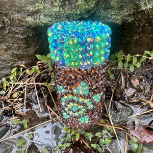 Puede incluir: Un vaso decorativo con un degradado de cristales iridiscentes azules, verdes y marrones. Los cristales cubren toda la superficie del vaso, creando un efecto brillante. El vaso está colocado al aire libre entre pequeñas plantas y rocas.