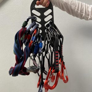 May include: A black climbing gear organiser holds various carabiners and quickdraws. The carabiners are in red, blue, and silver, with black and blue webbing. The organiser is held by a person.