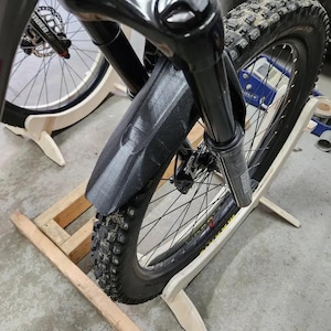 May include: A black mountain bike with a front suspension fork is mounted on a wooden bike stand. The bike has a black fender and a black tire with a tread pattern.