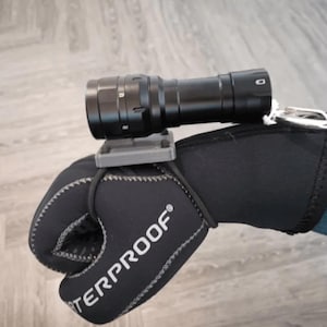 May include: A black flashlight mounted on a gray 3D printed mount attached to a black neoprene glove with the word "WATERPROOF" printed on it.