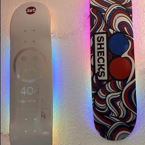 Può includere: Due skateboard appesi a un muro. La tavola a sinistra è bianca con un design a faccina triste, il testo "404 Page not found" e "Go back home". La tavola a destra ha un design a vortice con il testo "SHECKS".