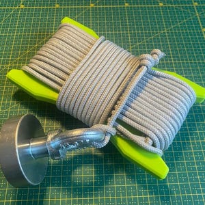 May include: A gray rope is coiled around a bright green spool, secured with a knot. A metal handle is attached to the spool. The items are on a green grid surface. This item is likely used for crafts or DIY projects.