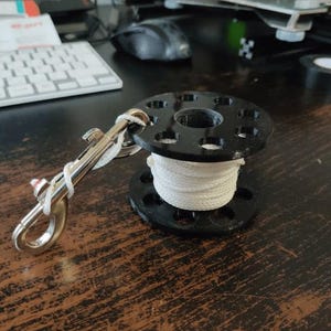 Könnte beinhalten: Eine schwarze Tauchrolle mit weißem Seil und einem silbernen Karabiner. Die Rolle hat ein kreisförmiges Design mit mehreren Löchern und liegt auf einer Holzoberfläche. Der Karabiner ist mit weißer Schnur an der Rolle befestigt.
