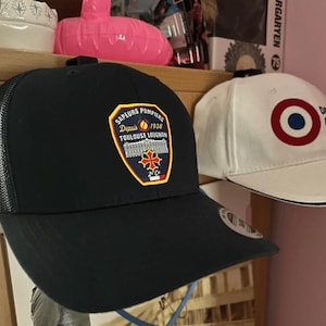 May include: Two baseball caps. The first is black with a mesh back and a patch that reads "Sapeurs Pompiers Depuis 1938 Toulouse Lougnon". The second is white with a red, white, and blue target design.