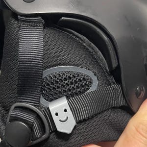 May include: Black helmet with a gray strap and a small gray sticker with a smiley face on it.