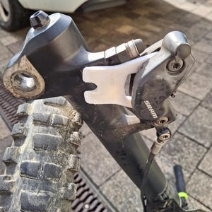 Peut inclure: Gros plan sur un étrier de frein de vélo noir avec un protège-étrier en plastique blanc. Le levier de frein est fixé au guidon et le câble de frein est visible. Le frein est marqué "SRAM".