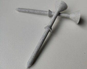 May include: Two golf tees with a speckled, light gray appearance. Each tee has a pointed end and a small, circular collar near the top. The tee heads are a flared, cup-like shape. The tees are lying on a white surface.