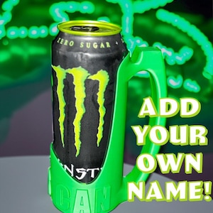 May include: A black and green Monster Energy drink can with a green handle. The can has the words "ZERO SUGAR" and the Monster logo in yellow. The green handle has the words "ADD YOUR OWN NAME!" printed on it.