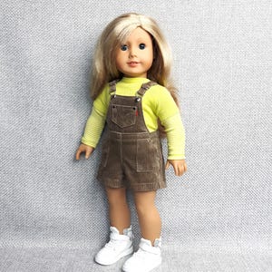 May include: A doll with blonde hair and blue eyes, dressed in a yellow long-sleeved top and brown corduroy dungaree shorts. The doll is wearing white high-top trainers.