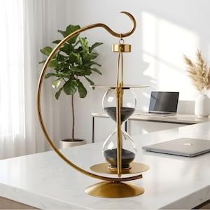 May include: A gold-colored hourglass with black sand, suspended within a curved metal frame. The hourglass features a circular base and a hook at the top. The background includes a white countertop, a laptop, and a potted plant.