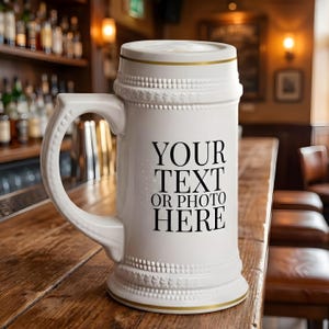 May include: White ceramic beer stein with a large handle and gold trim. The stein has the text "YOUR TEXT OR PHOTO HERE" printed in black. The stein is sitting on a wooden surface.