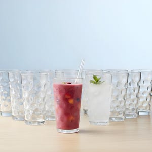 May include: A collection of clear drinking glasses with a textured, bubble-like pattern. Two glasses are filled with colorful beverages, one a red smoothie with fruit and the other a clear drink with ice and a mint sprig. A clear straw is in one glass.
