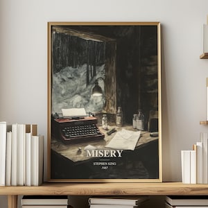 May include: A framed vertical art print featuring a dark, moody illustration of a vintage typewriter on a wooden desk, with a snowy window view, medicine bottles, and papers. The text reads MISERY, STEPHEN KING, 1987 in a clean white font.