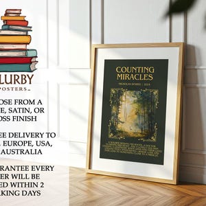 May include: A framed poster with the title "Counting Miracles" by Nicholas Sparks. The poster features a forest scene with a deer. The frame is light brown. The poster is displayed on a wooden surface.