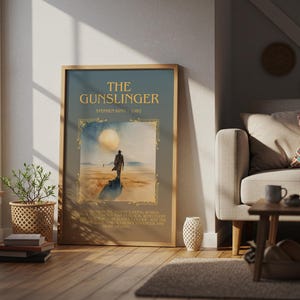 May include: Framed poster of "The Gunslinger" by Stephen King. The artwork depicts a lone figure in a desert landscape under a large moon. The poster is in a wooden frame and is displayed in a living room setting.