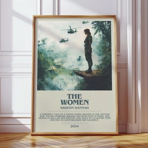 Puede incluir: Un p&oacute;ster enmarcado con el t&iacute;tulo "THE WOMEN" de Kristin Hannah. La imagen muestra a una mujer con uniforme militar de pie en una plataforma de madera, con helic&oacute;pteros al fondo. El p&oacute;ster est&aacute; sobre un suelo de madera.