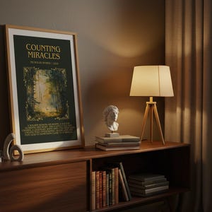 May include: A framed art print titled "Counting Miracles" with a forest scene. The print is displayed on a wooden cabinet, alongside a lamp, books, and a bust sculpture. The room has a warm, inviting atmosphere.