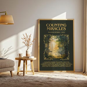 May include: A framed art print with the title "COUNTING MIRACLES" by Nicholas Sparks. The artwork features a forest scene with a deer and text at the bottom. The frame is gold.