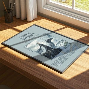 May include: A framed print titled "Onyx Storm" on a wooden desk. The artwork features a fantasy scene with dragons and mountains. The text includes the words "Violet faces rising threats" and "She battles dark magic".