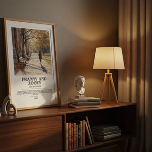 K&ouml;nnte beinhalten: Gerahmtes Poster mit dem Text "FRANNY AND ZOOEY" von J.D. Salinger, das eine Person zeigt, die auf einem von B&auml;umen ges&auml;umten Weg geht. Eine Holzlampe mit beigem Schirm und eine B&uuml;stenskulptur stehen auf einem Holzregal.