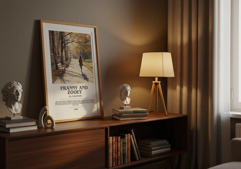 K&ouml;nnte beinhalten: Gerahmtes Poster mit dem Text "FRANNY AND ZOOEY" von J.D. Salinger, das eine Person zeigt, die auf einem von B&auml;umen ges&auml;umten Weg geht. Eine Holzlampe mit beigem Schirm und eine B&uuml;stenskulptur stehen auf einem Holzregal.