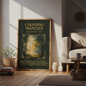 May include: A framed poster with the title "COUNTING MIRACLES" by Nicholas Sparks. The poster features a forest scene with a deer. The text is in gold on a dark green background. The poster is displayed in a living room setting.