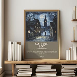 May include: A framed poster for Stephen King's "Salem's Lot" featuring a dark, atmospheric illustration of a town at night with a full moon. The poster has a gold frame and is displayed on a wooden shelf with books.