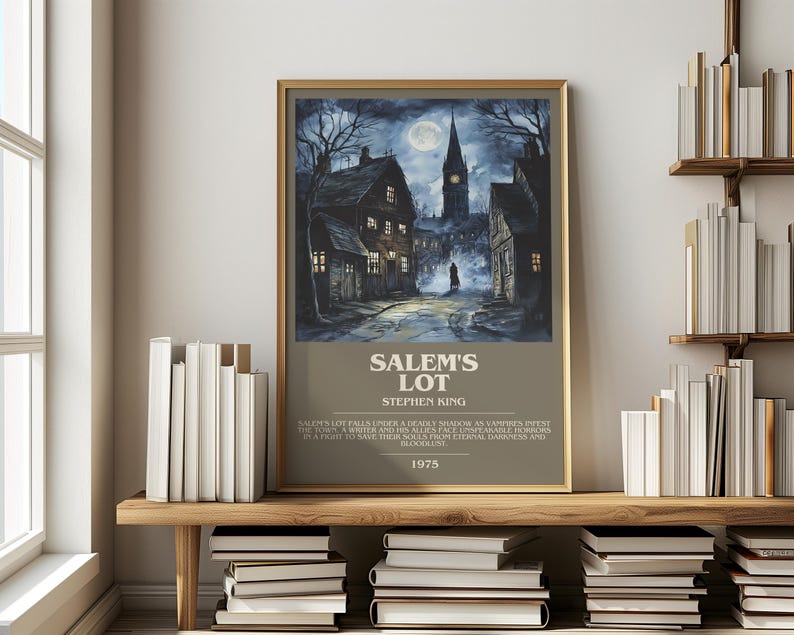 May include: A framed poster for Stephen King's "Salem's Lot" featuring a dark, atmospheric illustration of a town at night with a full moon. The poster has a gold frame and is displayed on a wooden shelf with books.