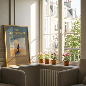 May include: A framed poster titled "The Gunslinger" leans against a wall next to a window. The poster features a figure on a beach with a large sun. Small potted plants sit on the windowsill. Two armchairs are in the foreground.