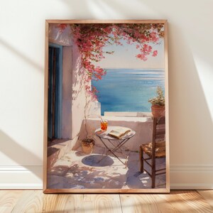 Puede incluir: Impresión enmarcada de una serena escena de balcón con vistas al mar. La obra representa una pared blanca con una puerta azul, una pequeña mesa con una bebida y un libro, y una silla de madera. Flores rosas caen desde arriba, añadiendo color.