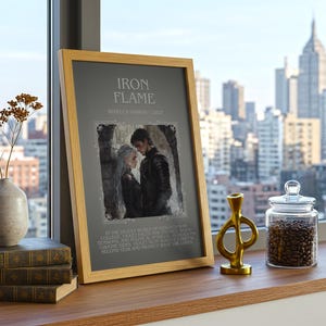 May include: A framed print with the text "IRON FLAME" and an illustration of a couple. The print is displayed on a wooden surface with books, a vase with dried flowers, a gold-colored sculpture, and a glass jar filled with coffee beans. The background shows a cityscape.