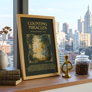May include: A framed print titled "Counting Miracles" by Nicholas Sparks, displayed on a wooden surface. The print features a forest scene and text. Beside it are books, a vase with dried flowers, a gold sculpture, and a glass jar filled with coffee beans.