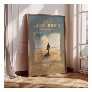 May include: A framed art print of "The Gunslinger" by Stephen King. The artwork depicts a desert landscape with a lone figure, a large moon, and gold text. The frame is gold, and the colour scheme is muted, giving a vintage feel.
