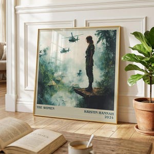 May include: A framed art print featuring a woman in military attire overlooking a jungle scene with helicopters. The print has a gold frame and includes the text "THE WOMEN" and "KRISTIN HANNAH 2024".