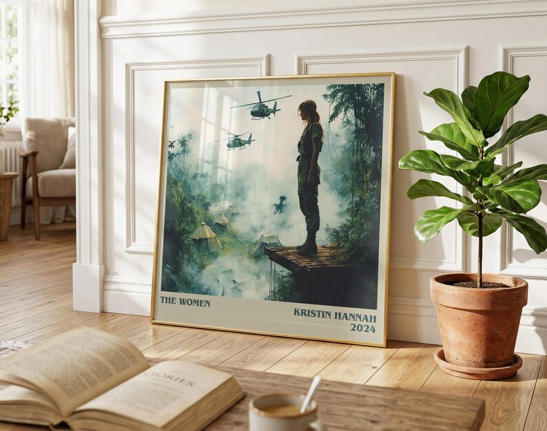 May include: A framed art print featuring a woman in military attire overlooking a jungle scene with helicopters. The print has a gold frame and includes the text "THE WOMEN" and "KRISTIN HANNAH 2024".