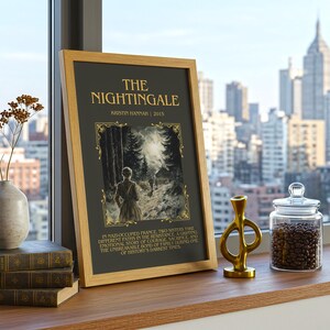 Peut inclure: Une impression encadr&eacute;e de "The Nightingale" de Kristin Hannah, expos&eacute;e sur une surface en bois avec des livres, un vase et un objet d&eacute;coratif dor&eacute;. Un bocal en verre rempli de grains de caf&eacute; est &agrave; proximit&eacute;, avec une vue sur la ville en arri&egrave;re-plan.