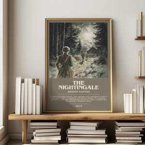May include: A framed print of "The Nightingale" by Kristin Hannah, featuring a woman walking on a path through a forest. The print has a gold frame and is displayed on a wooden shelf with books.